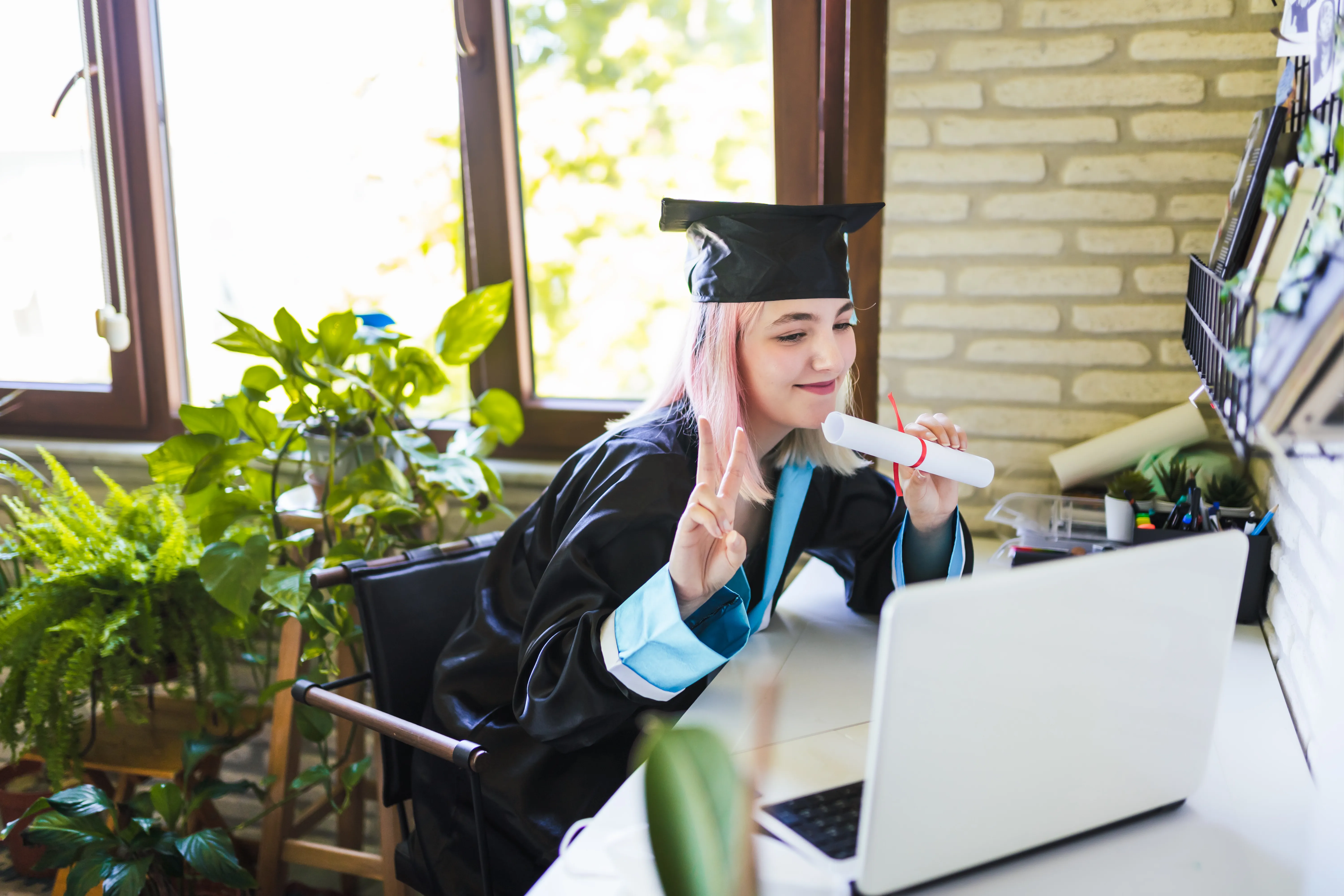 Graduate in cap and gown making a peace sign during a video call on a laptop.