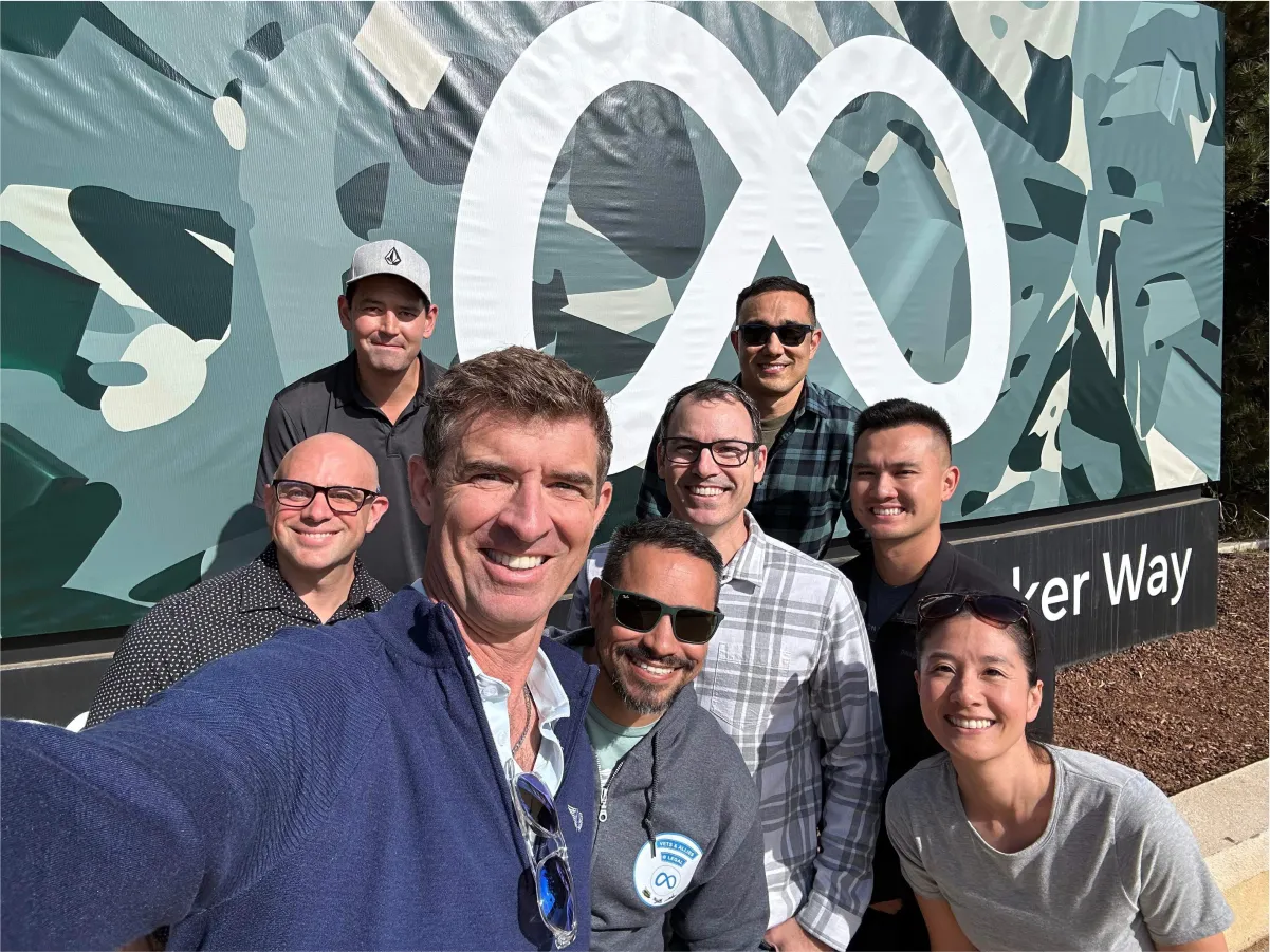 Seven adults smile for a selfie in front of a camouflage banner with a white infinity logo.