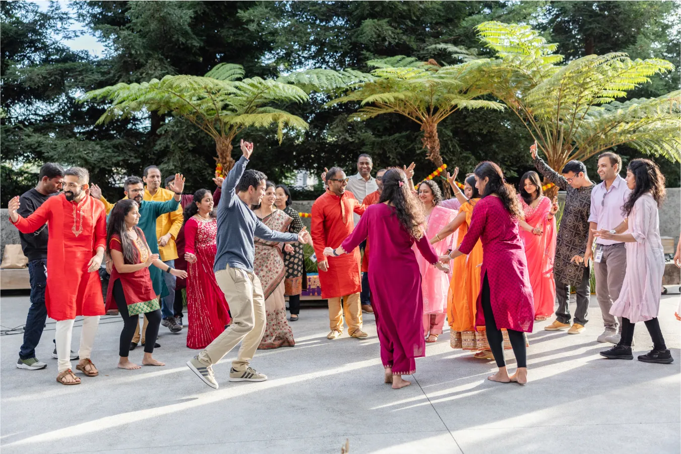 A group of people in colorful Indian attire dancing outdoors with festive decorations and trees in the background.