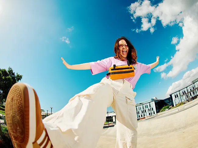 A teen standing on one leg with their arms out smiles for a photo outside on a sunny day.