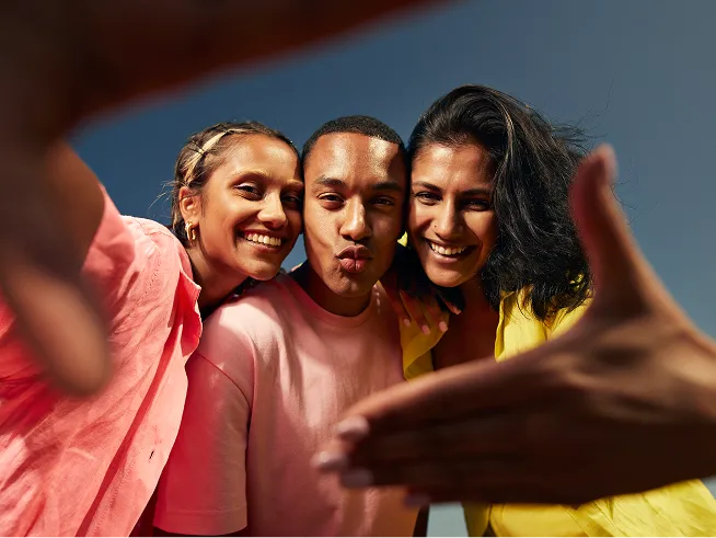 Three young adults pose for a joyful selfie together, holding their hands up to create a frame around their faces.
