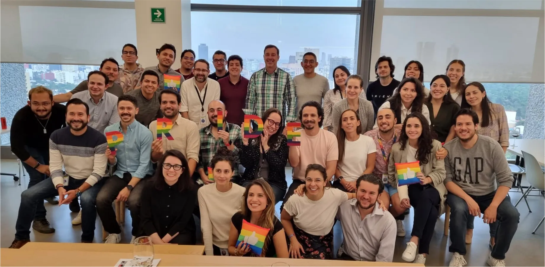 A group of coworkers pose in an office holding rainbow “PRIDE” letters and other rainbow items to celebrate Pride.