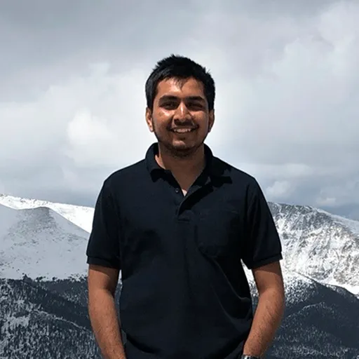 A man in a black polo shirt standing in front of snow‑capped mountains.
