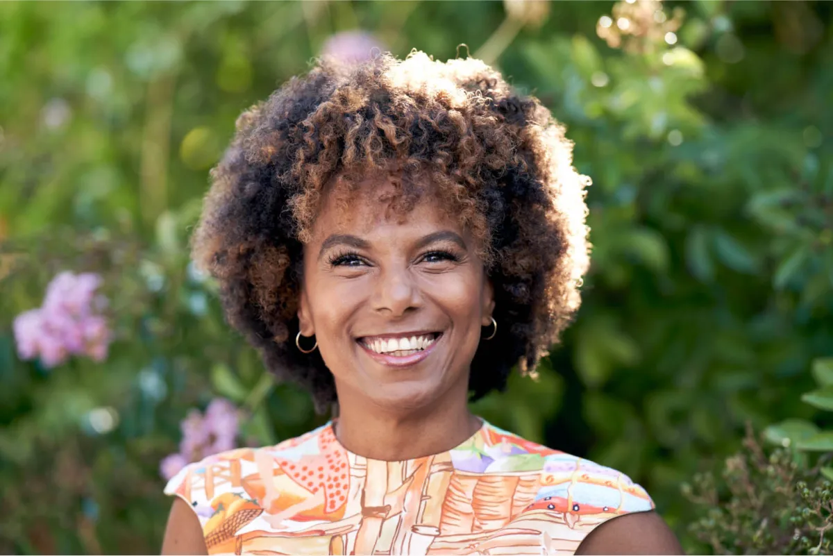 A woman with curly brown hair smiles outdoors, wearing a colorful patterned sleeveless top and gold hoop earrings.