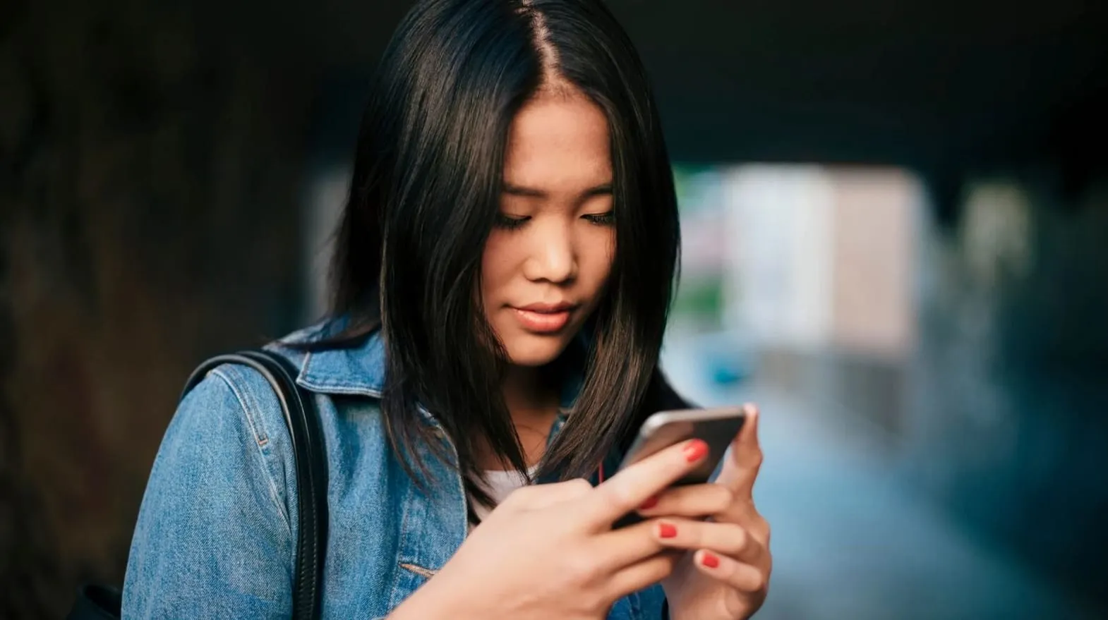 Teen focused on a phone while standing outdoors, wearing a denim jacket and backpack.