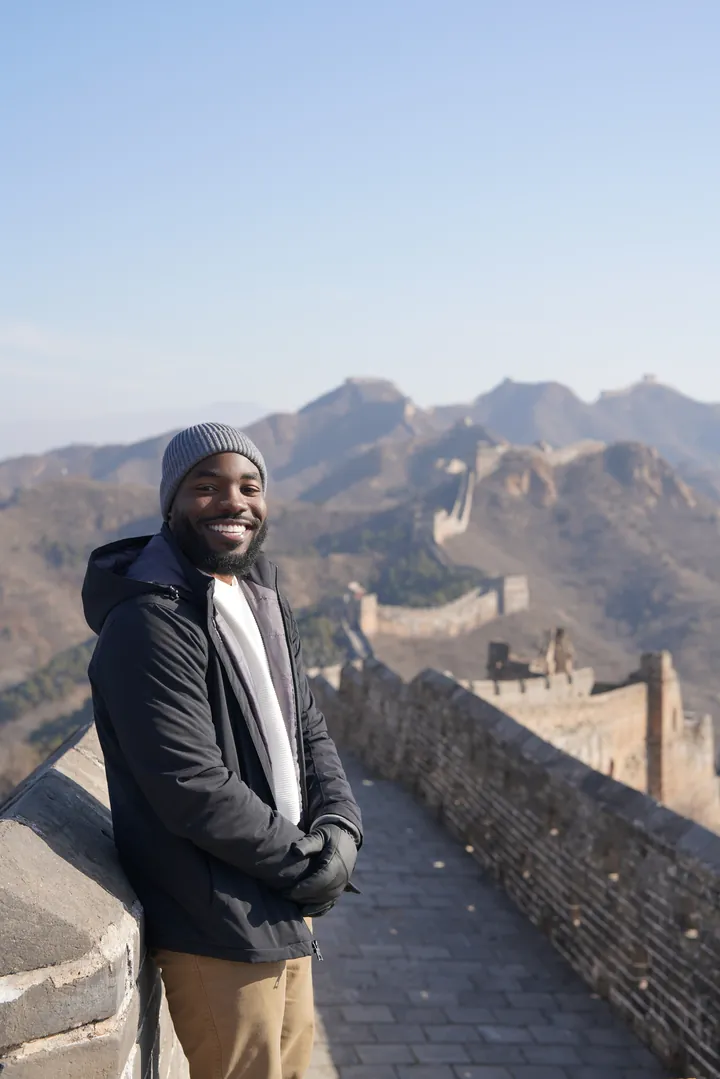Erekosima standing on the Great Wall of China wearing a beanie and black jacket, the wall winding through mountains behind him
