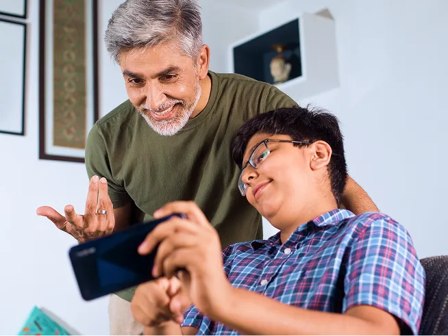 A teen points to his phone while his parent smiles and looks at the screen.