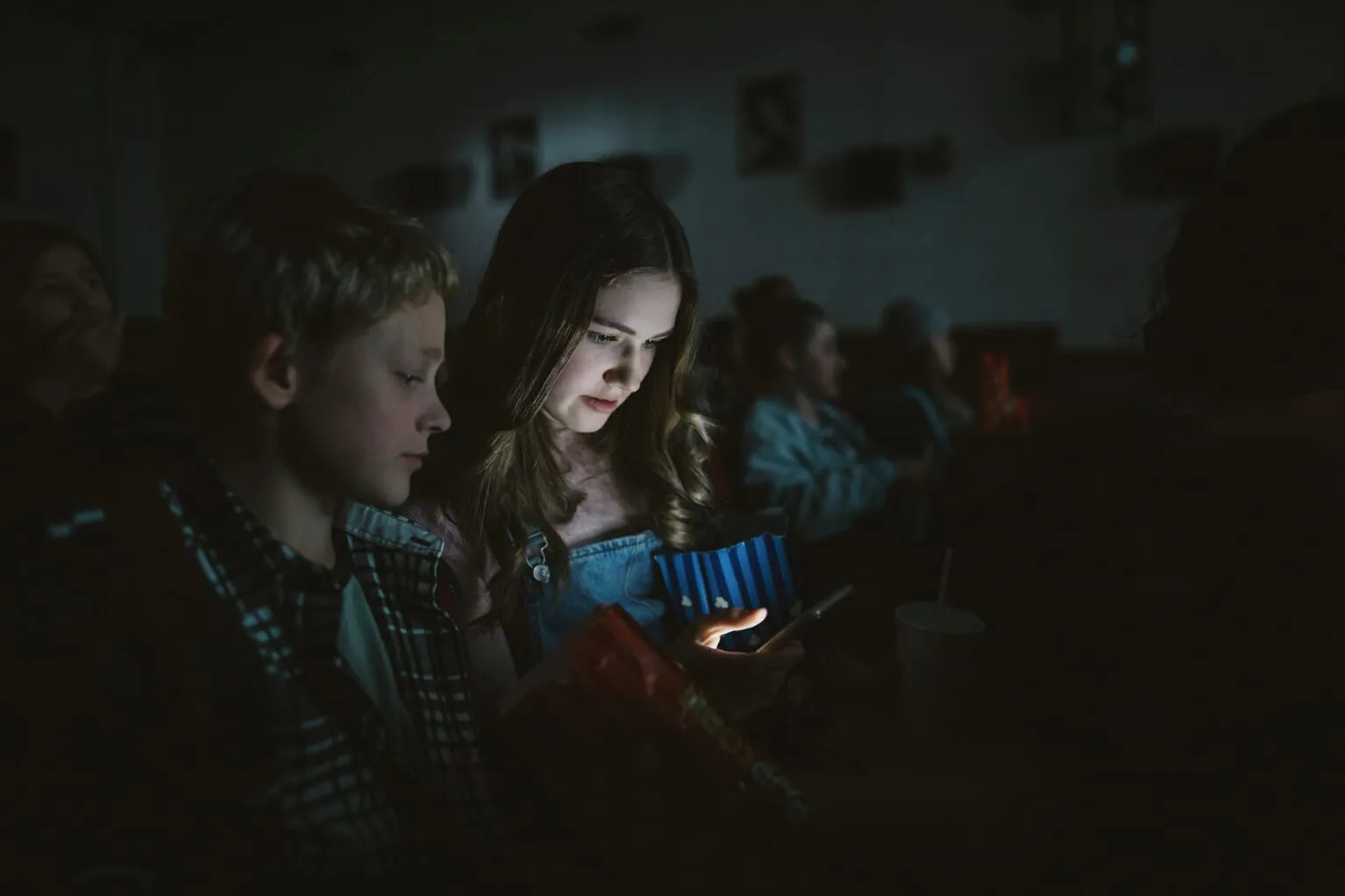 Duas pessoas sentadas em um cinema escuro, uma delas olhando para uma tela de smartphone brilhante.