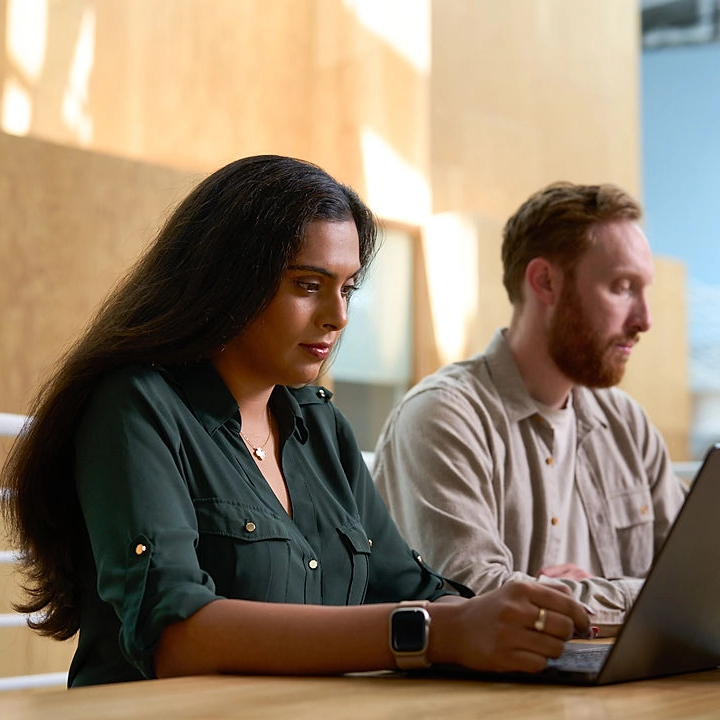 Woman in a green blouse types on a laptop next to a colleague in a sunlit office.