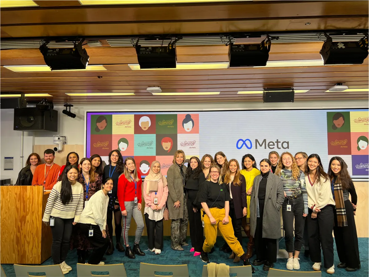 About 20 women pose for a photo in front of a large screen showing the Meta logo and a “Women @ Meta” illustration grid.