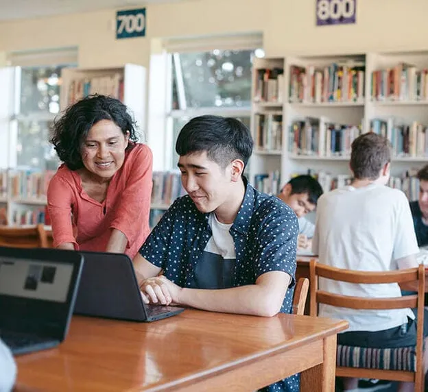 Teacher assisting a student working on a laptop in a library.