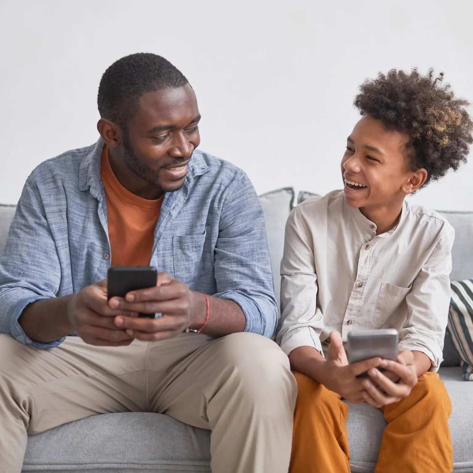 Un adulto e un bambino seduti su un divano che si sorridono mentre tengono in mano degli smartphone.