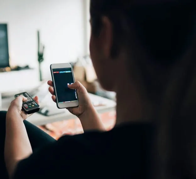 Person holding a phone and a remote, interacting with a screen in a home setting.
