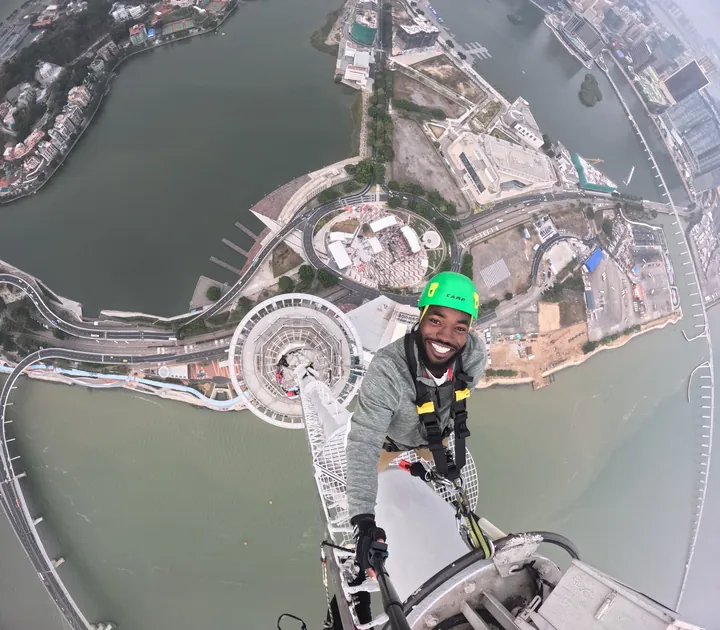 Erekosima grinning in a safety harness and green helmet at the top of Macau Tower with the city far below