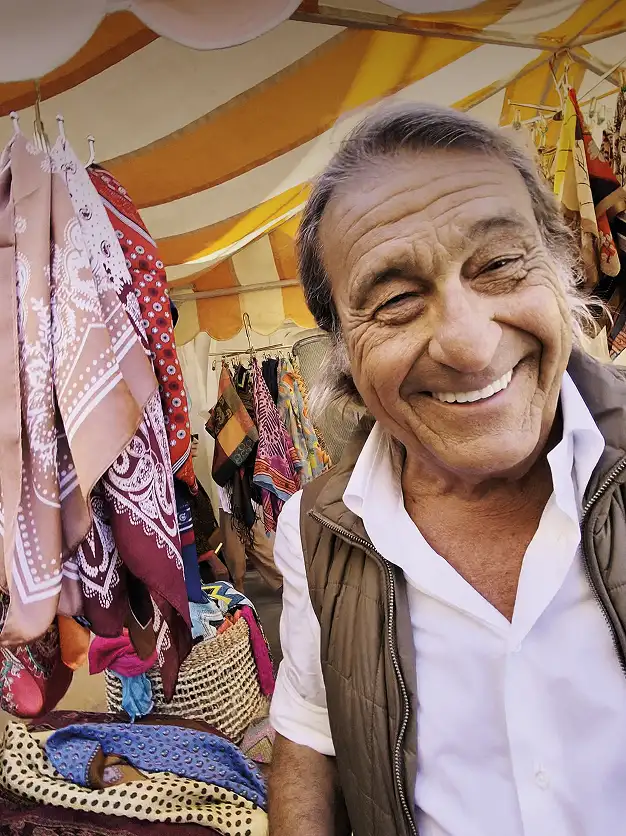 A smiling older man at an outdoor market surrounded by colorful scarves.