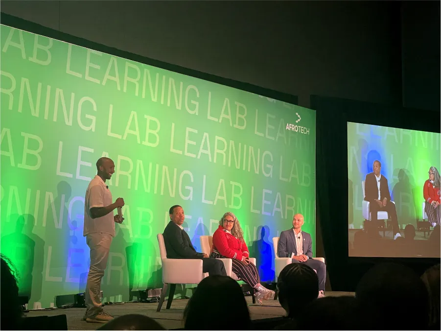 A stage with a green “LEARNING LAB” Afrotech backdrop shows three panelists seated while a host stands with a microphone.