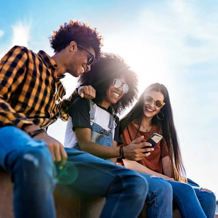 Three teens sitting outdoors in the sun, smiling and looking at a phone together.