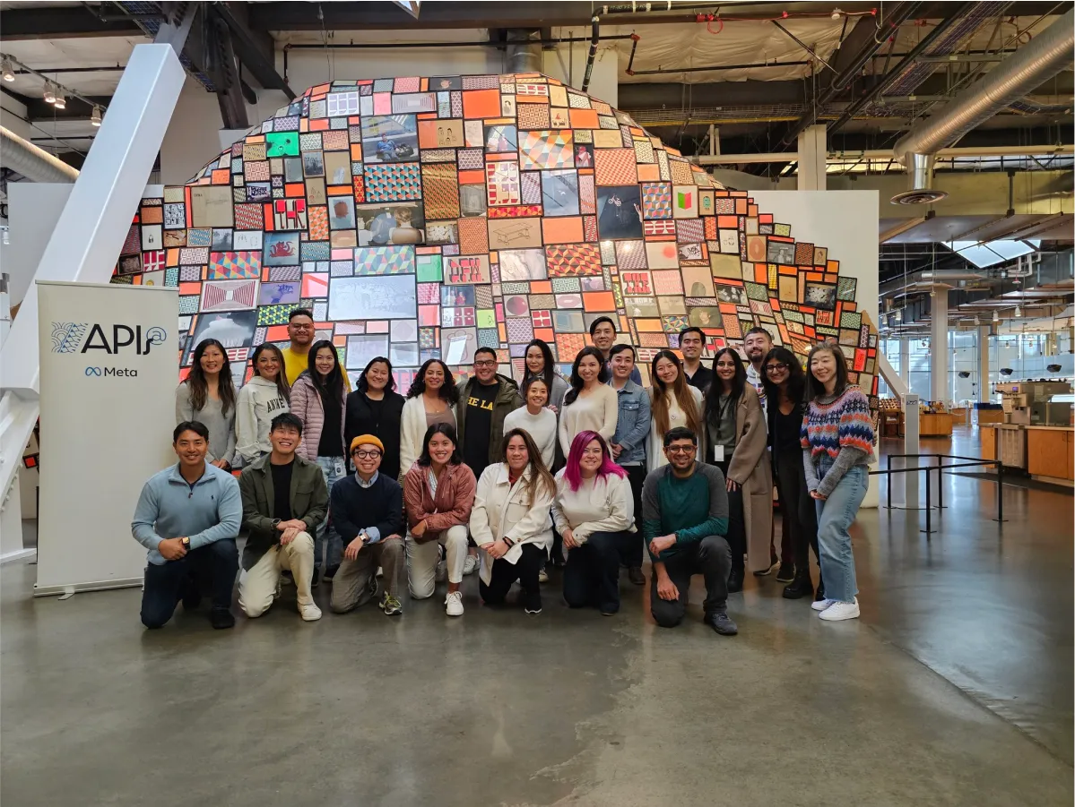 Twenty-two diverse people pose for a group photo in front of a large colorful patchwork dome installation inside an industrial indoor space.
