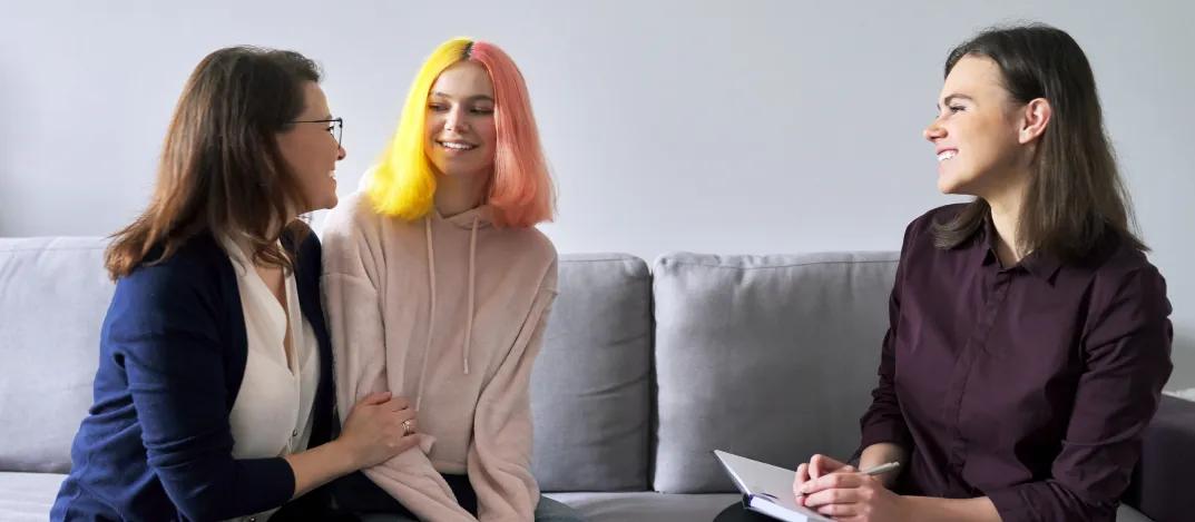 Une personne aidante, un jeune aux cheveux teints en jaune et rose, et un·e professionnel·le de la santé mentale assis ensemble sur un canapé, tous souriant lors d’une discussion bienveillante.