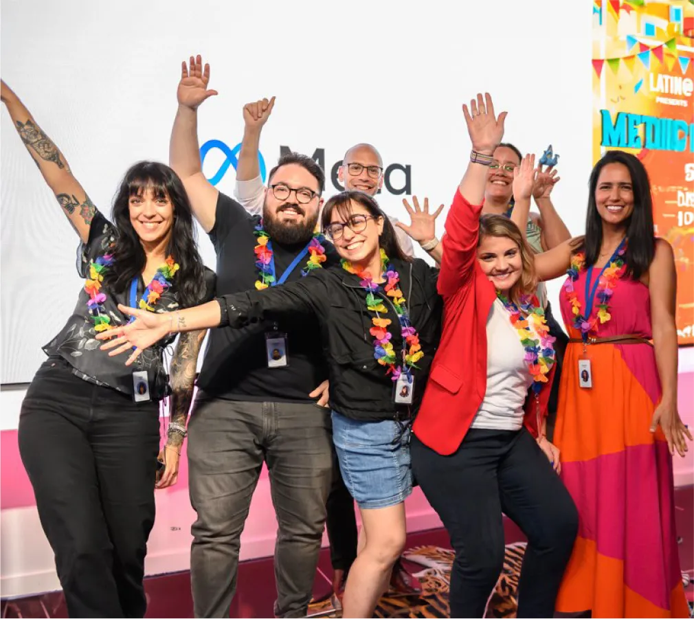 Six people wearing colorful flower leis and blue ID lanyards pose enthusiastically in front of a white backdrop with a partial Meta logo.