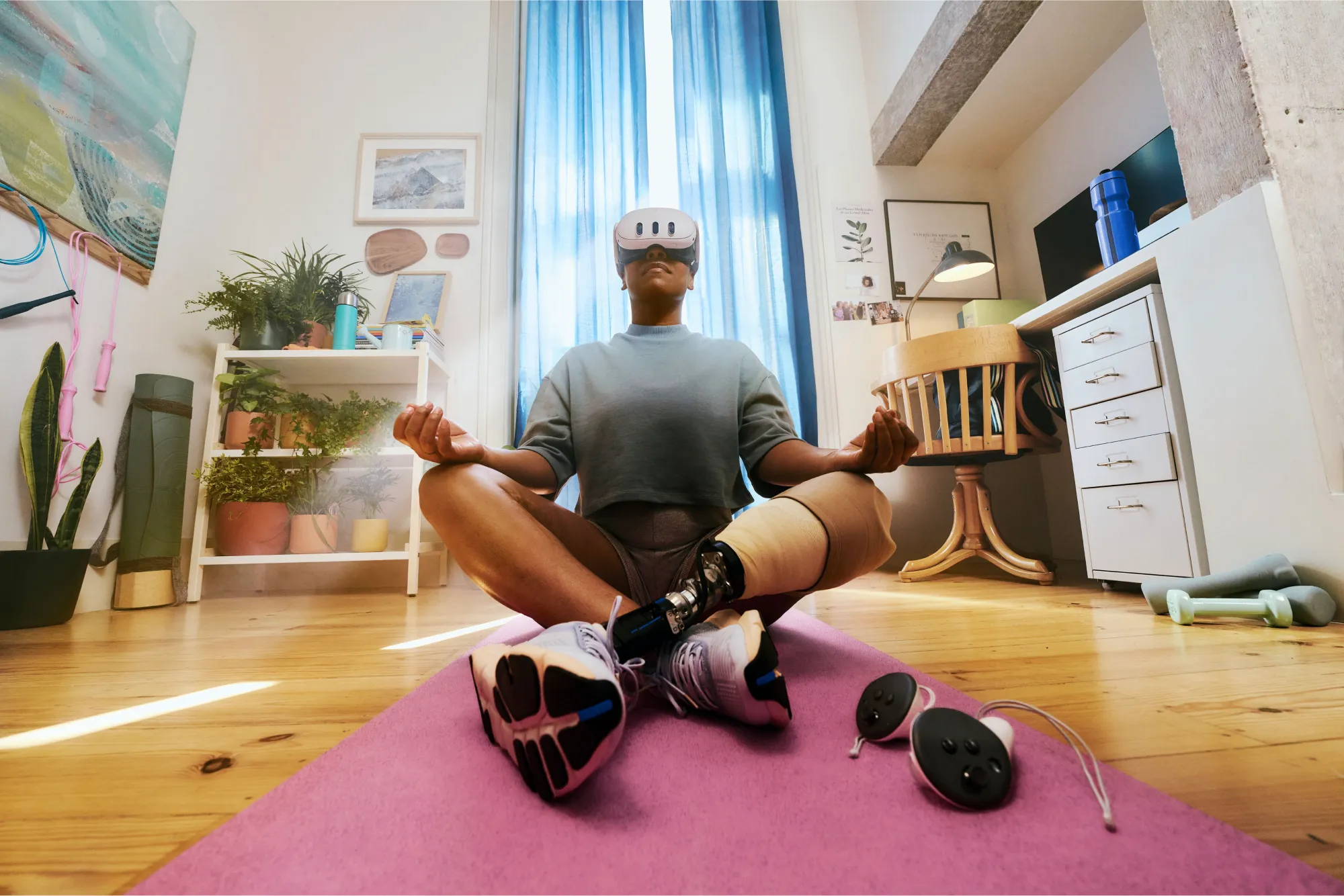 A person with a prosthetic leg wears a VR headset while sitting cross-legged in a meditation pose on a pink yoga mat in a cozy home.