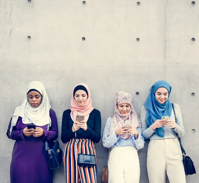 Four people wearing hijabs standing against a wall, each using a smartphone.