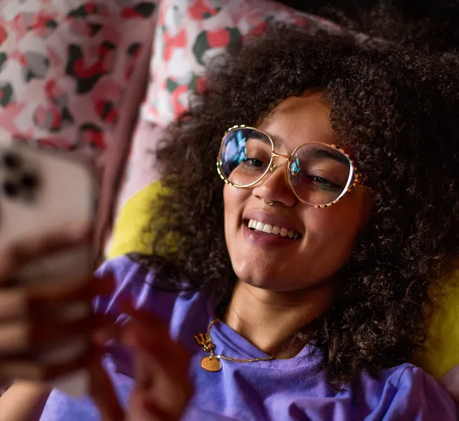 Gros plan d’un jeune aux cheveux bouclés et portant de grandes lunettes, souriant en regardant l’écran de son téléphone.