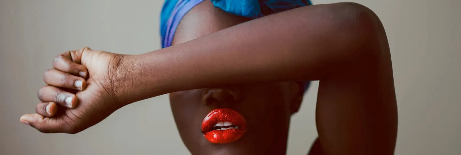 Primo piano di una persona con un rossetto acceso, che si copre parzialmente il viso con il braccio.