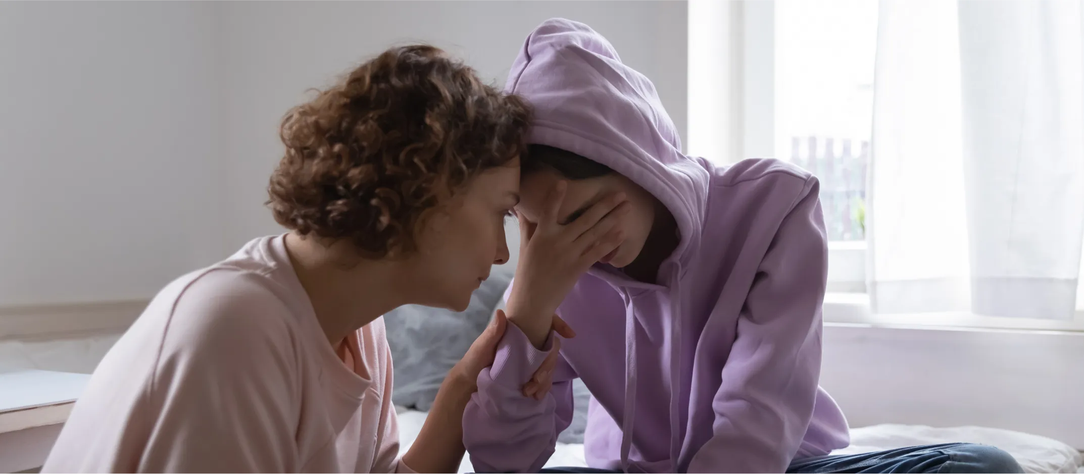 Une personne aidante réconforte un jeune en chandail à capuchon lavande, visiblement bouleversé, leurs fronts se touchant dans un moment d’empathie et de connexion.