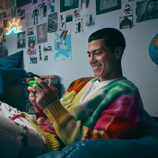 Person sitting on a couch smiling while playing a game on a phone, with colorful posters on the wall in the background.