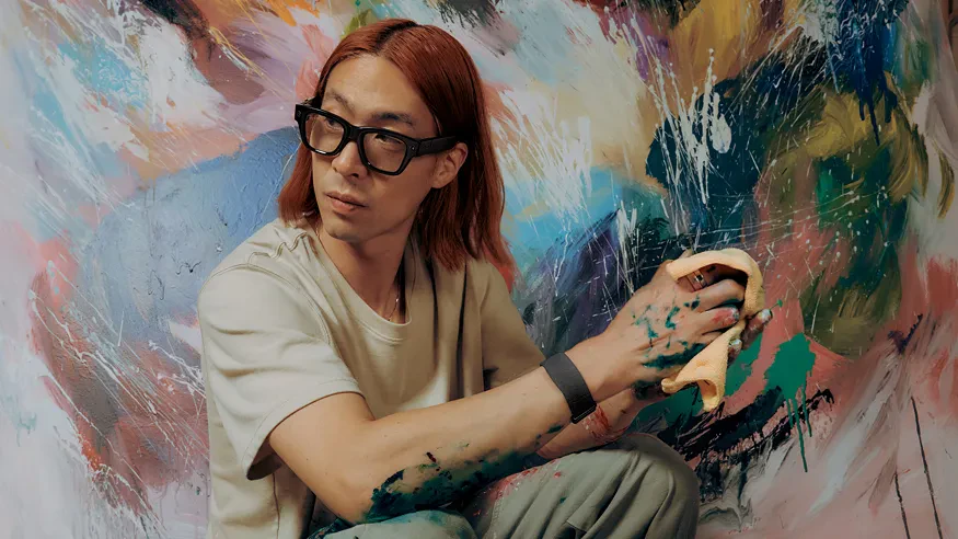 Man sits in front of abstract painting with cloth in hands wearing Meta Ray-Ban Display glasses.