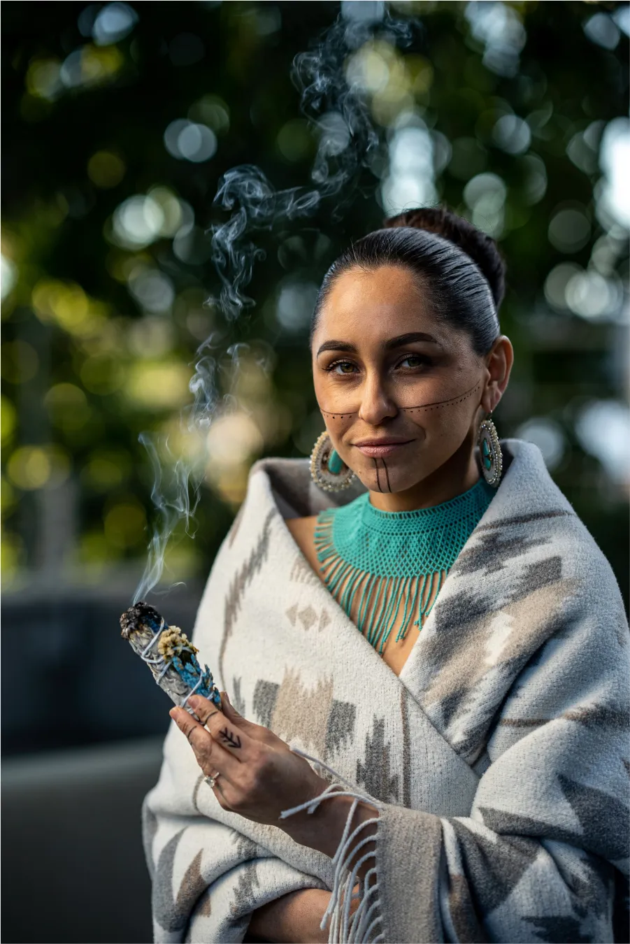 A woman holds a smoking sage bundle, wearing turquoise earrings and black facial markings.
