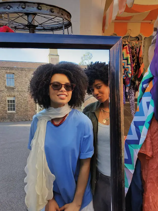 Two friends standing outside next to a mirror and a rack of colorful clothing, one wearing Meta glasses.