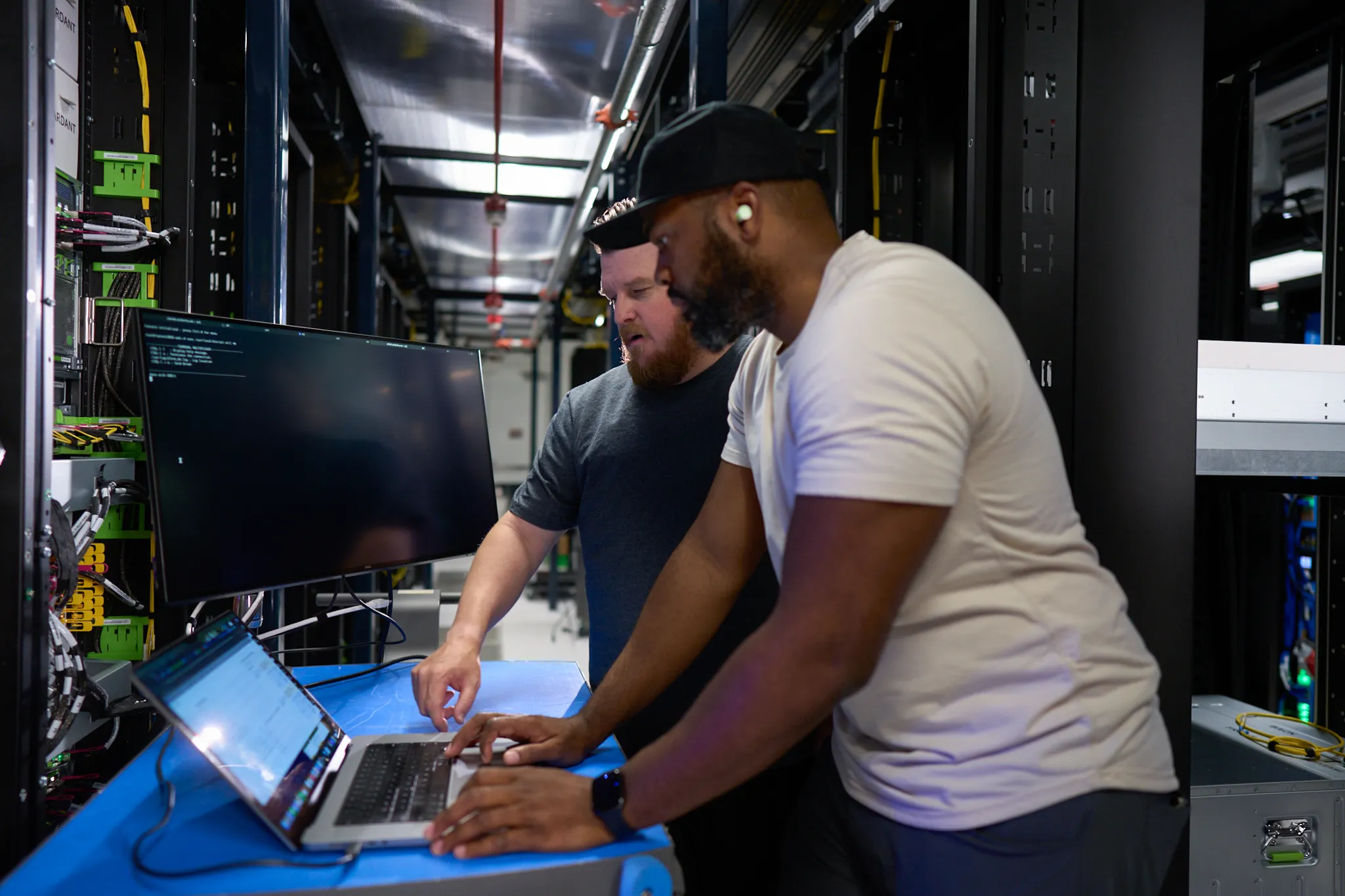Two men working on a laptop in a server room.