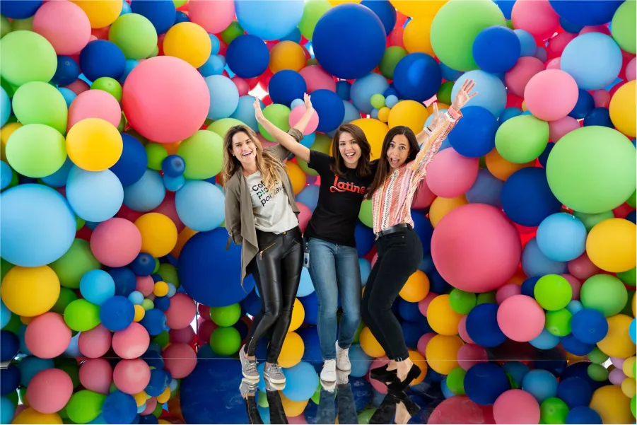 Three women jumping in front of a colorful balloon wall backdrop.