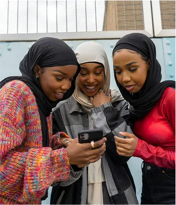 建物の前でスマートフォンを囲み、画面上で何かをシェアしながら微笑む3人のティーン。