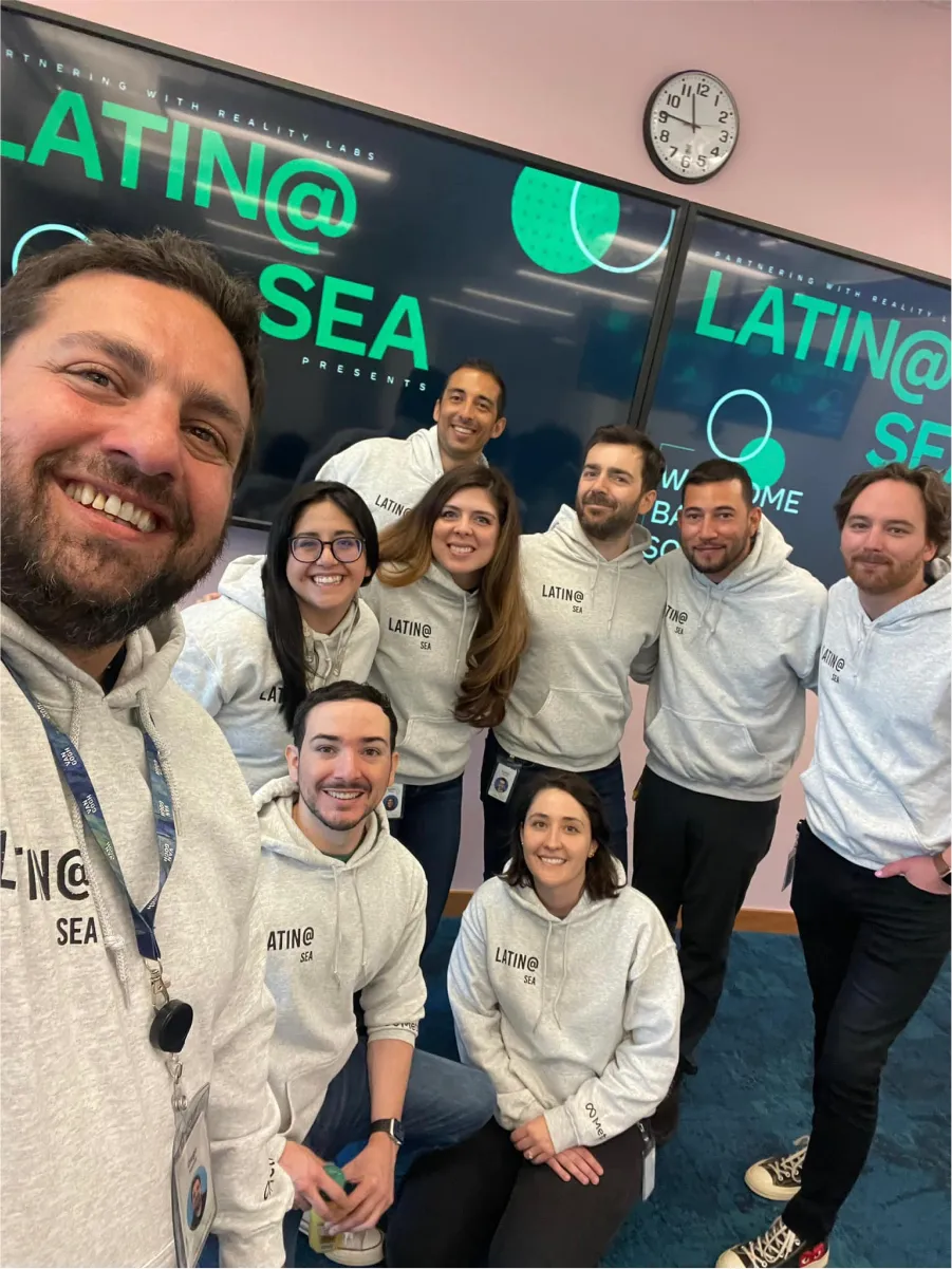 Eight people in matching “LATIN@ SEA” hoodies pose for a selfie in front of two large “LATIN@ SEA” screens.