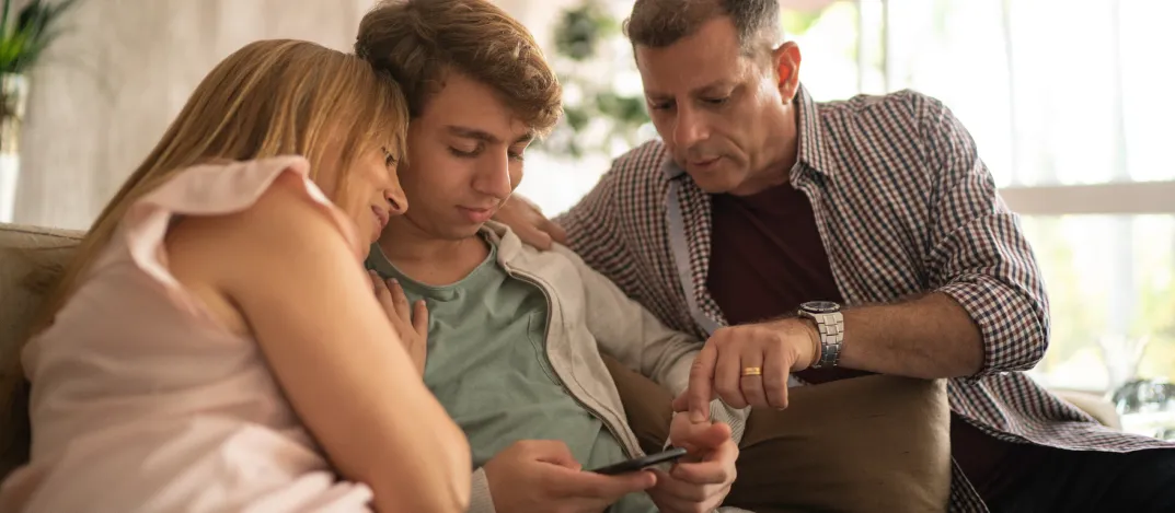 Deux parents et leur jeune assis côte à côte sur un canapé, regardant un téléphone ensemble et engagés dans une conversation.