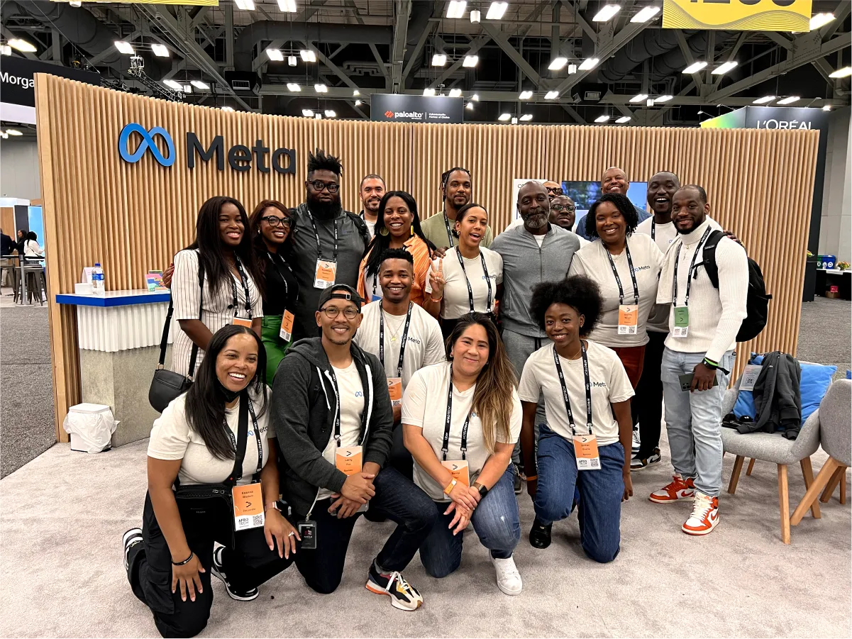 A group of 16 diverse adults posing for a photo at a Meta booth with wooden slat branding and orange event badges.