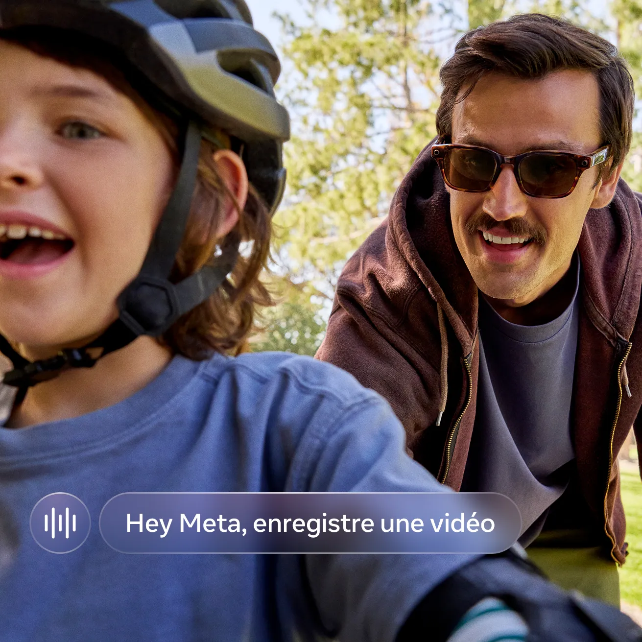 Un homme portant des lunettes Meta fait du vélo à côté d’un petit enfant souriant qui porte un casque sur un sentier bordé d’arbres.