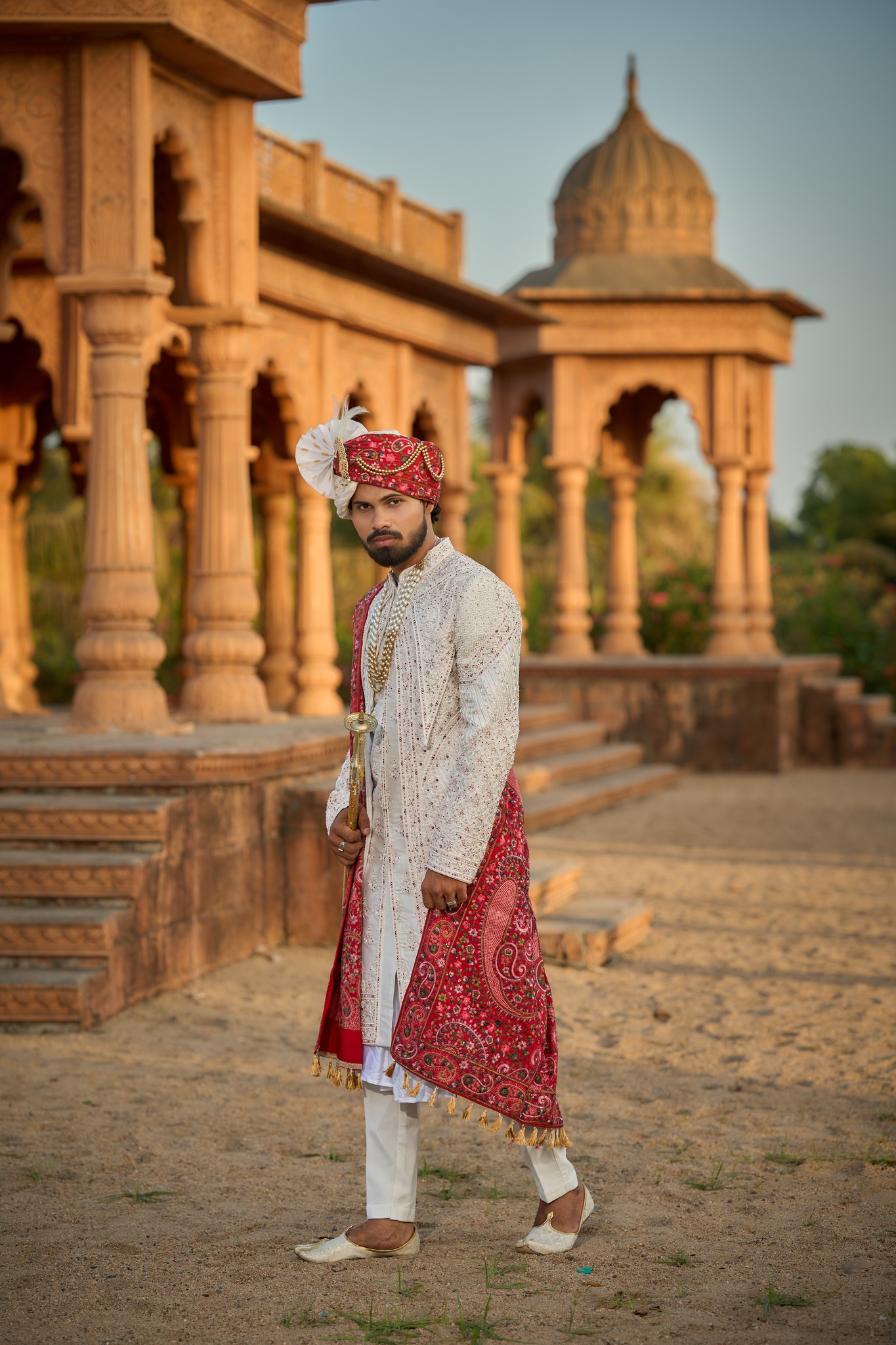 ✨ Royal Groom Look 👑, Ivory embroidered sherwani paired with a vibrant red paisley stole, traditional royal safa, layered pearl necklaces, and classic mojaris—crafted for the groom who carries 