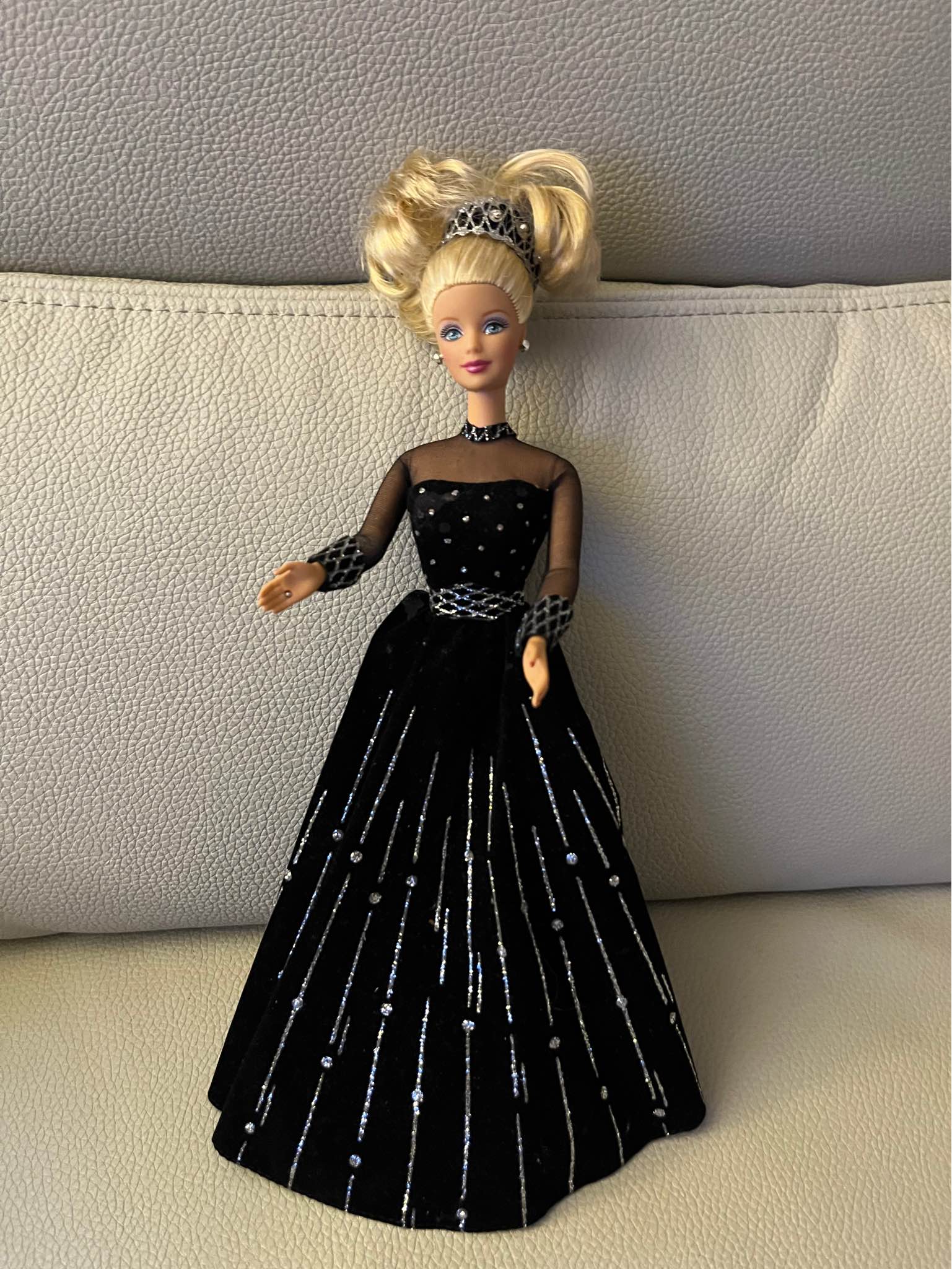 Elegant Barbie doll in a black velvet gown with silver glitter accents. The dress has sheer black mesh sleeves and a high neckline. The doll features blonde hair styled in a high