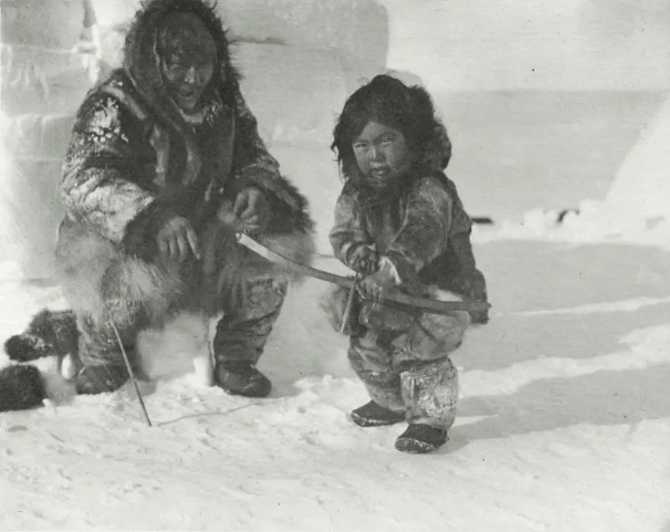 An Inuk toddler in full furs holding onto a sled, taken likely in