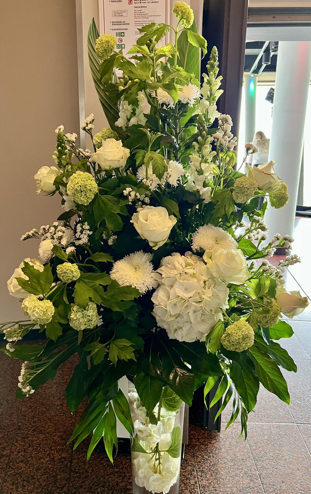 Ein üppiges Gesteck in edler Grün-Weiß-Komposition: Samtige Hortensien bilden das Herzstück, umrahmt von makellos weißen Rosen. Filigrane Chrysanthemen und elegante Löwenmäulchen verleihen Struktur und Höhe, während sattes Blattwerk in harmonischen