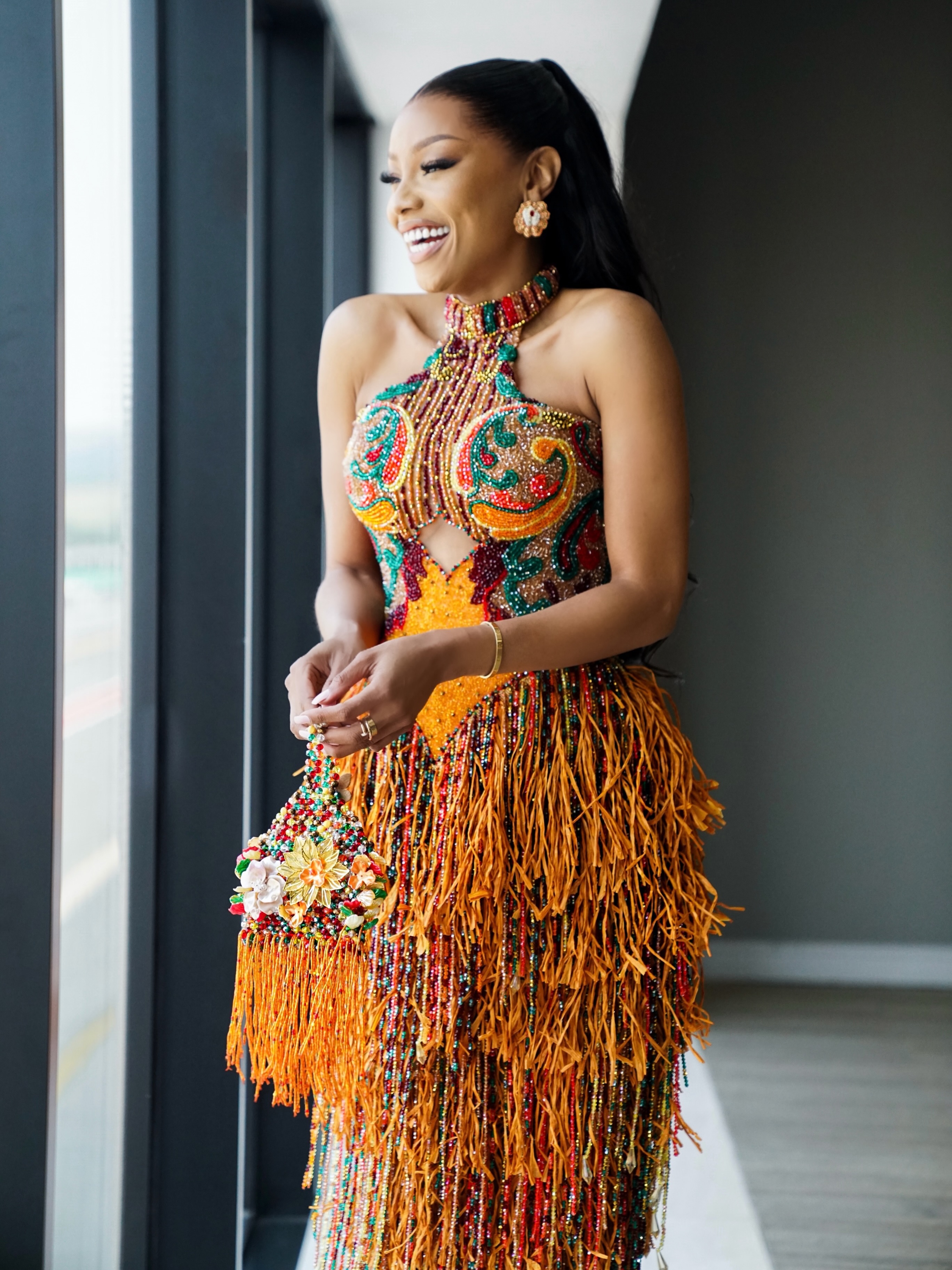 BonangMatheba looks stunning in this richly embellished orange, green and gold beaded gown by Nigerian designer, EmaginebyBukola. The halter-neck design features intricate beadwork, a sculpted bodice and dramatic cascading fringe, styled with
