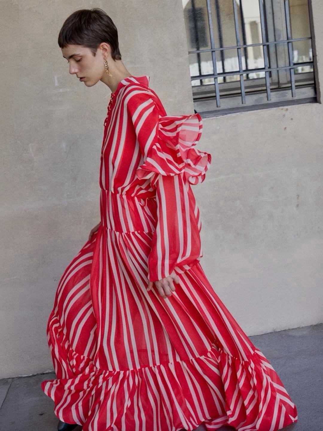 Long dress in coral and cream striped fabric, defined by a fluid silhouette and graphic rhythm. The deep V-neckline, ruffled sleeves and gathered waist create a soft structure, while the wide skirt