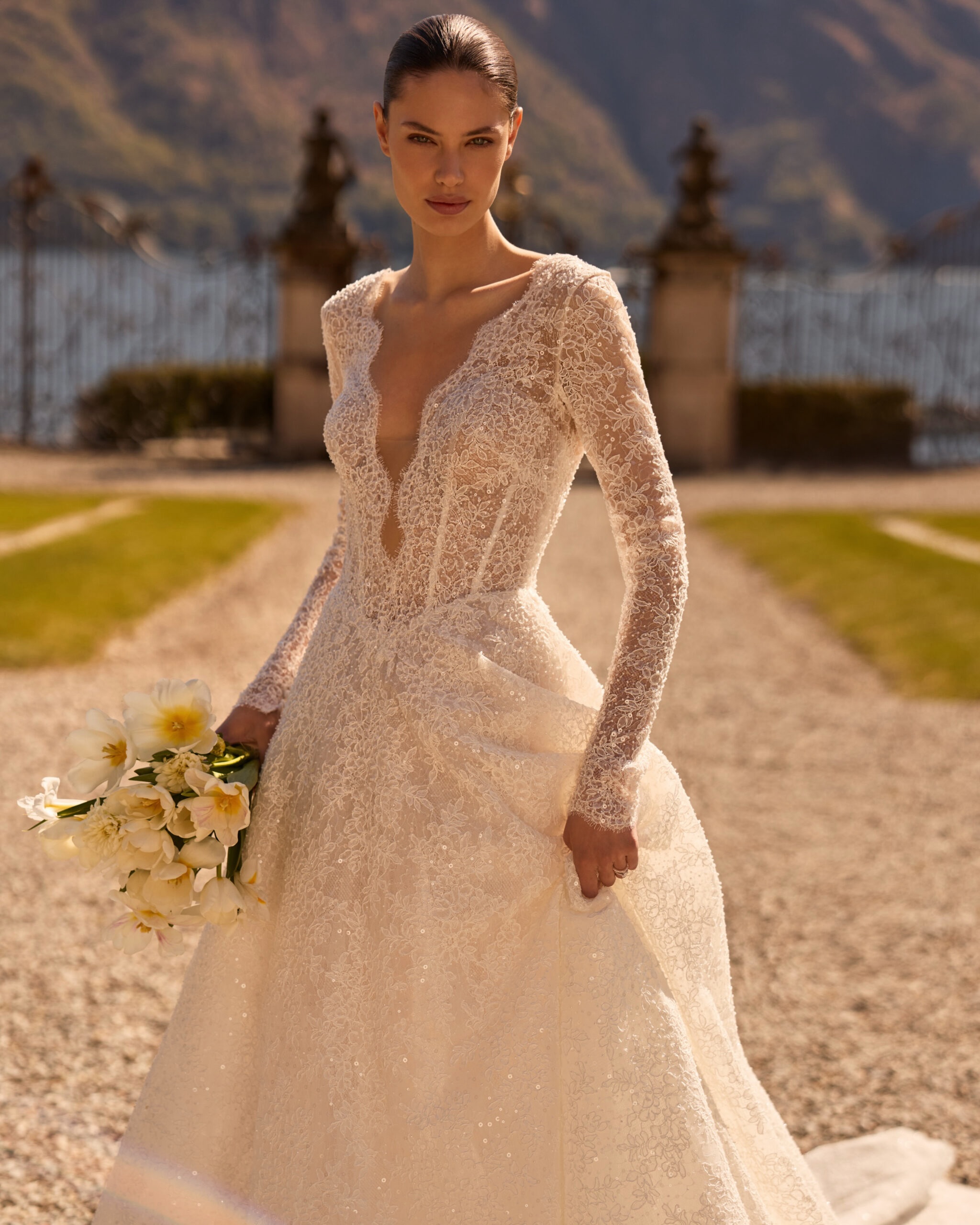 A classic princess-cut wedding dress crafted from lace with beading. It features a deep V-neck and long sleeves made from the same fabric. The V-back flows into a full, voluminous skirt that