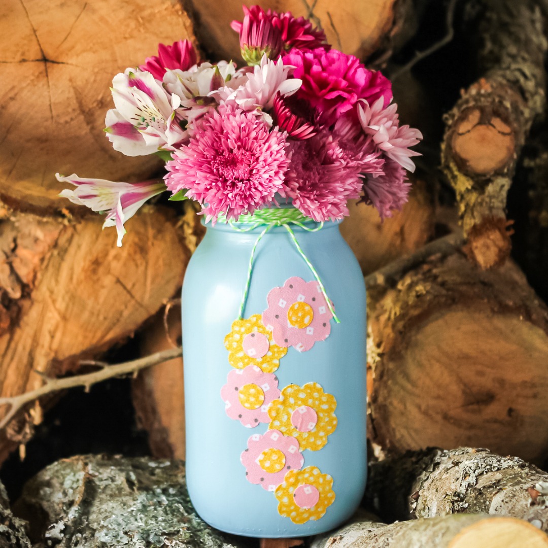 I love the fabric flowers adorning the front of this painted mason jar vase! 🌸 Grab your Cricut machine and get ready to make this spring craft idea in minutes. You can