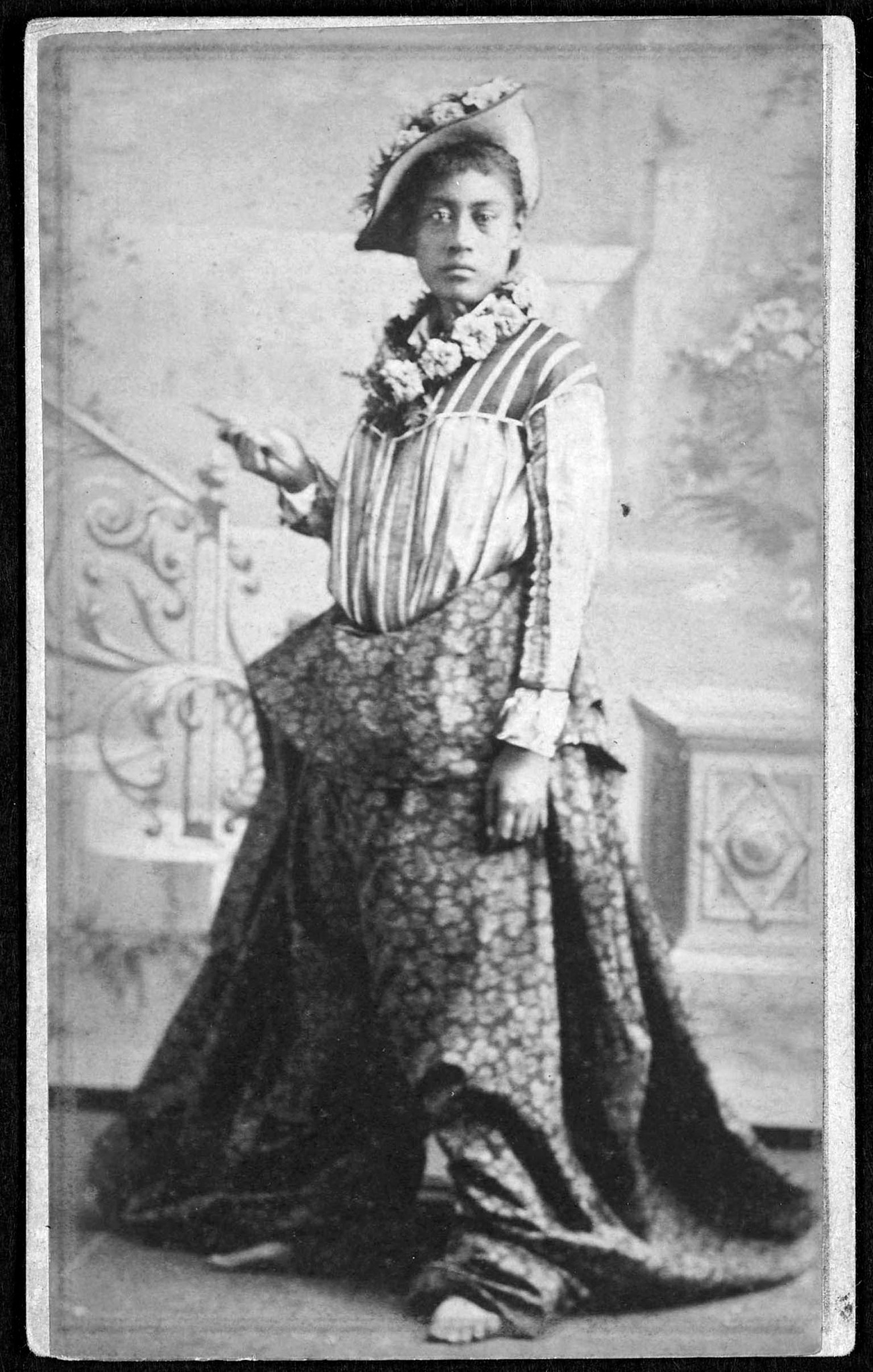 An undated studio portrait of a young Hawaiian woman in pāʻū riding attire, photographed by Andreas Avelino Montano. Her lei, hat, and flowing garments were carefully arranged for the camera, but her