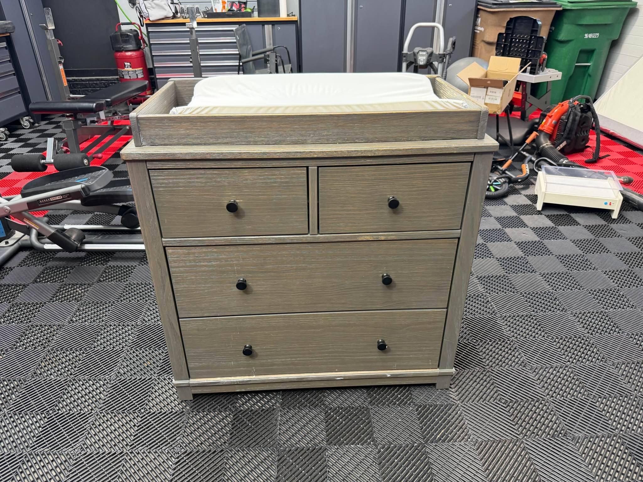 rustic grey dresser with changing table Gray wood nursery has four drawers black round knobs. The piece is constructed from wood with a weathered gray finish. It includes a removable topper and a white Diaper changing